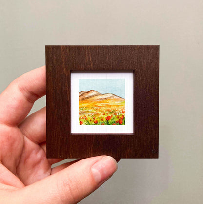 A hand is holding a small, square-shaped wooden frame containing a painting of a field with mountains in the background.