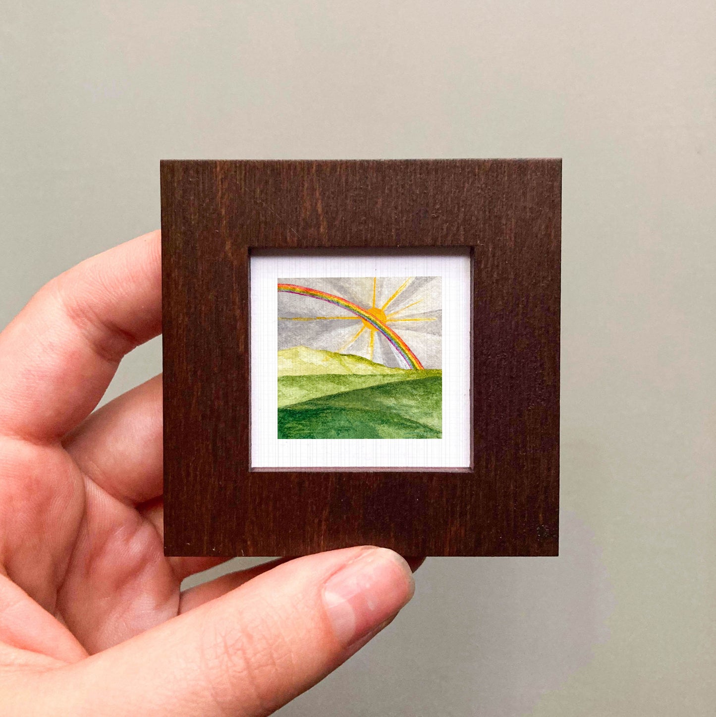 A hand is holding a small, square-shaped wooden frame containing a watercolor painting of a landscape with a rainbow in the background.