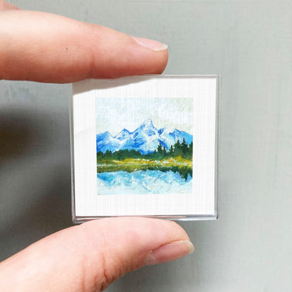 A person's hand holding a small square picture of a mountain landscape with a lake and trees.