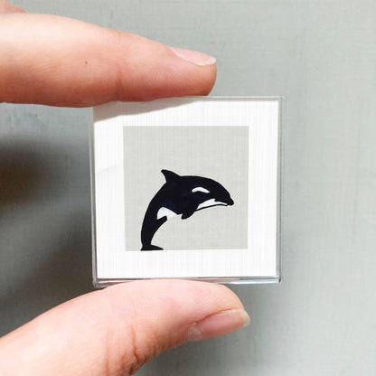 A person's hand is holding a small square with a black and white image of a dolphin on it.