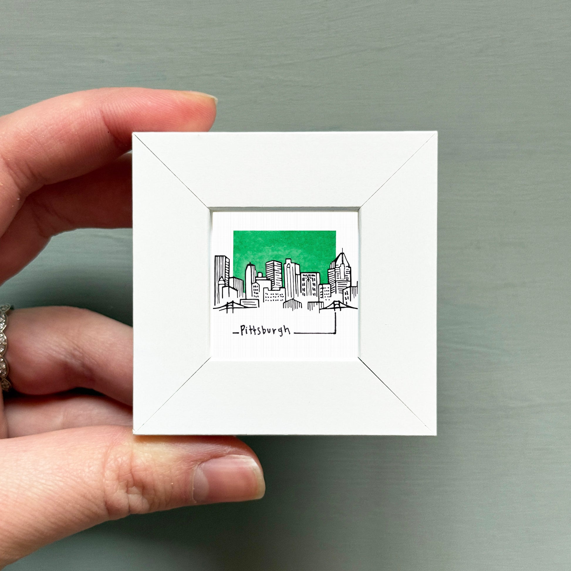 A hand is holding a small white square frame containing a black and white illustration of a city skyline.