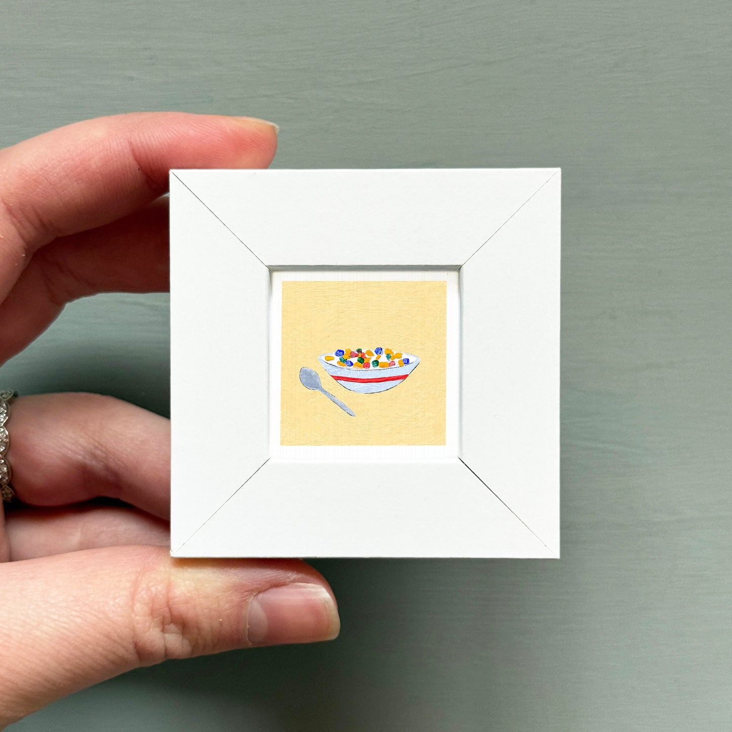 A hand is holding a small white picture frame containing an illustration of a bowl of cereal.