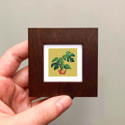 A hand is holding a small wooden frame containing a framed illustration of a potted plant with large leaves.