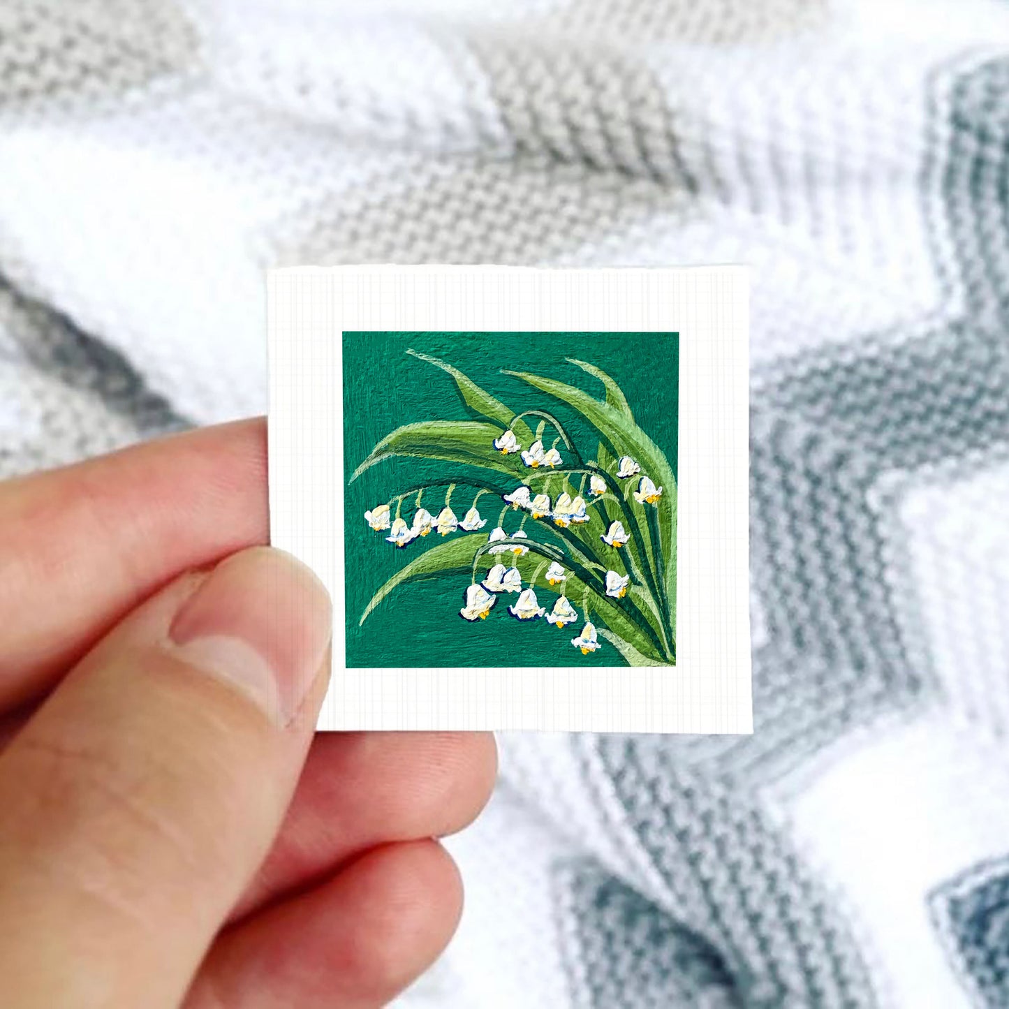 A hand is holding a small square card with a painting of lily of the valley flowers on it.