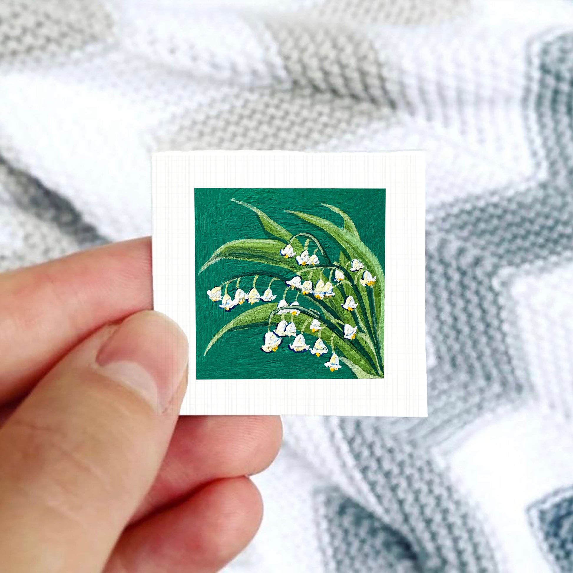 A hand is holding a small square card with a painting of lily of the valley flowers on it.