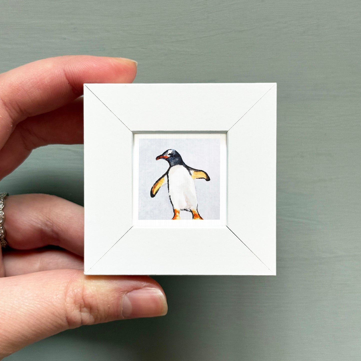 A person's hand is holding a small white square frame containing a picture of a penguin.