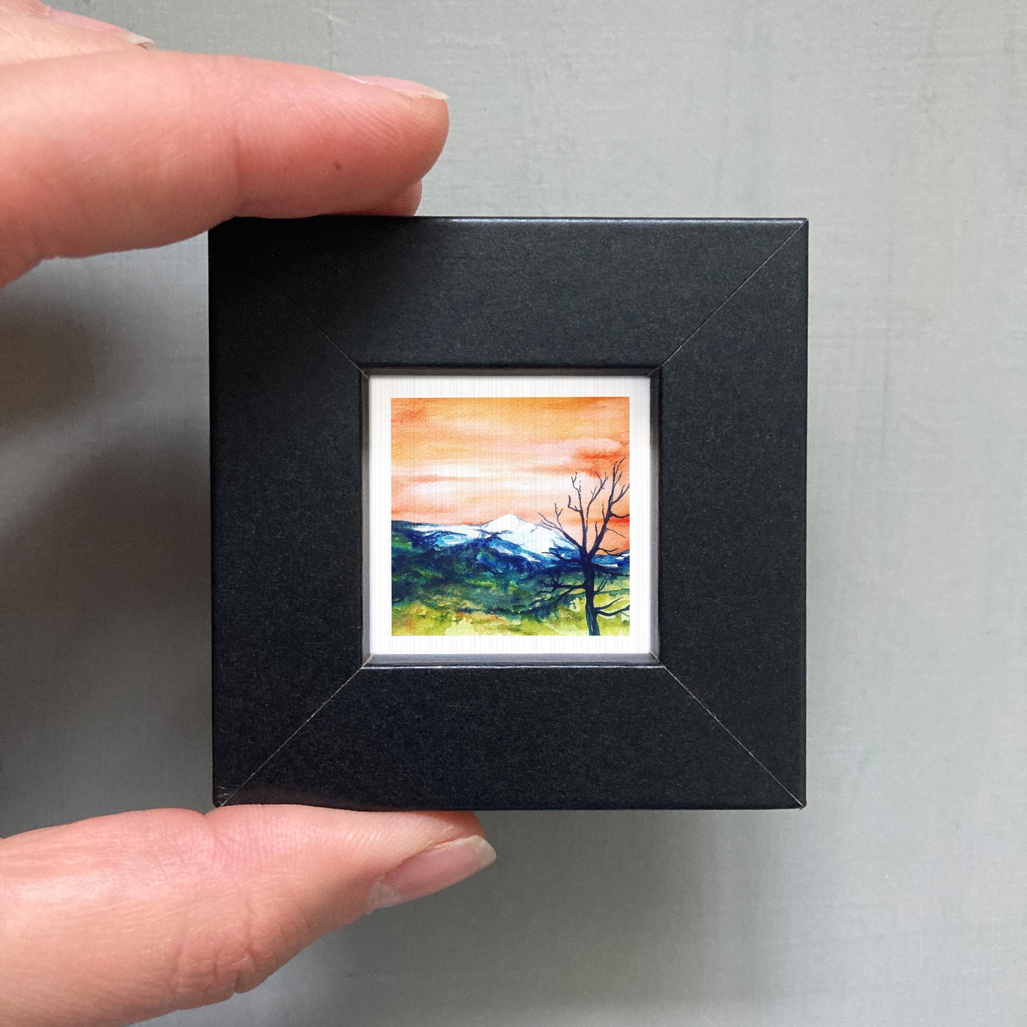 A person's hand is holding a small, square-shaped black frame containing a watercolor painting of a mountain landscape with a tree in the foreground.