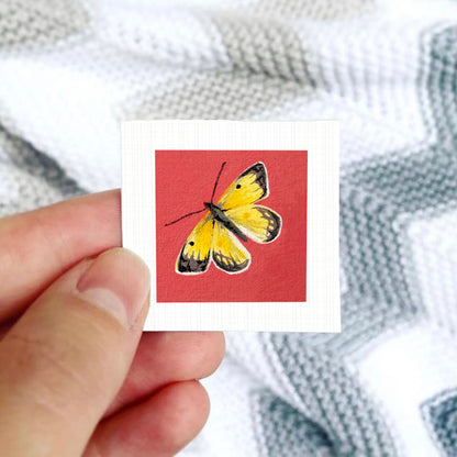 A hand is holding a small square card with a vibrant painting of a yellow butterfly on a red background.