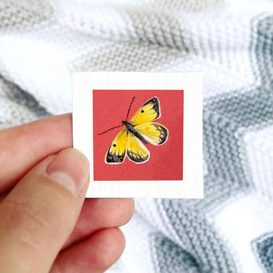 A hand is holding a small square card with a vibrant painting of a yellow butterfly on a red background.