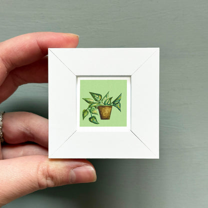 A hand is holding a small white square frame containing a painting of a potted plant.