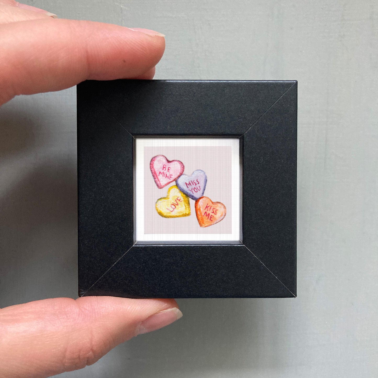 A hand is holding a small black picture frame containing a colorful image of four heart-shaped candies with messages on them.