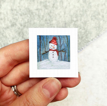 A hand holding a small square picture of a snowman against a snowy background.