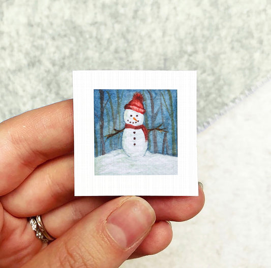A hand holding a small square picture of a snowman against a snowy background.