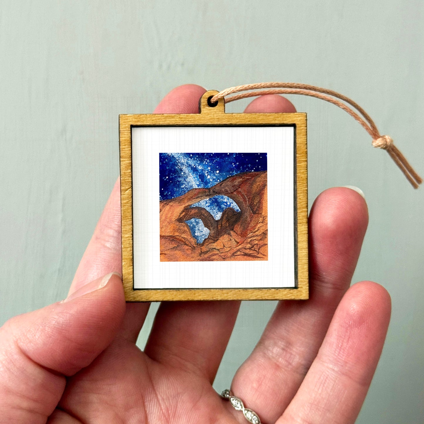 A hand holding a small wooden frame containing a painting of a desert landscape with a starry night sky.