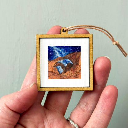 A hand holding a small wooden frame containing a painting of a desert landscape with a starry night sky.