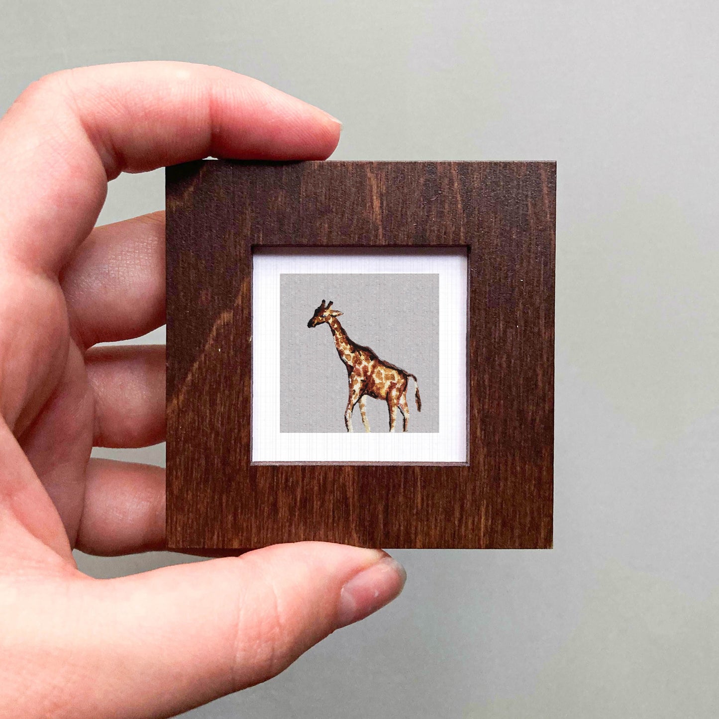 A hand is holding a small wooden frame containing a framed image of a giraffe.
