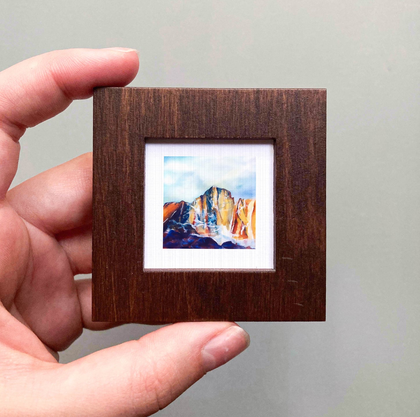 A hand is holding a small wooden frame containing a colorful painting of a mountain landscape.