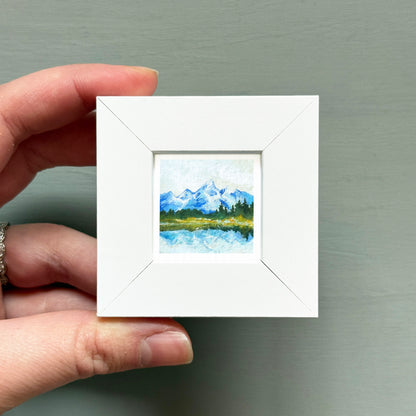 A hand is holding a small white square frame containing a painting of a mountain landscape.