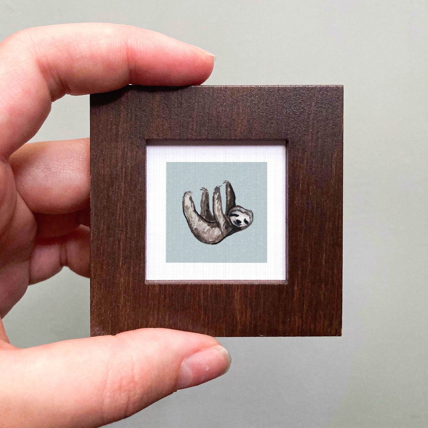 A hand is holding a small wooden frame containing a picture of a sloth hanging upside down.
