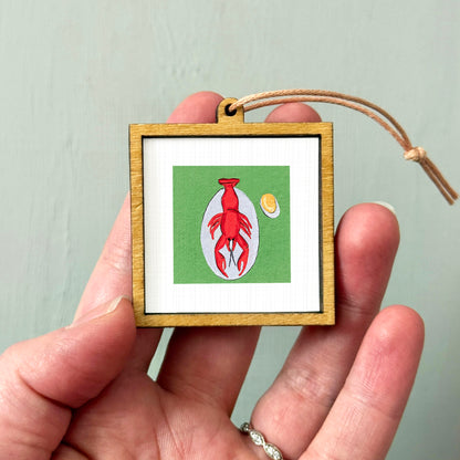 A person's hand holding a small wooden frame containing a picture of a red lobster and an egg.