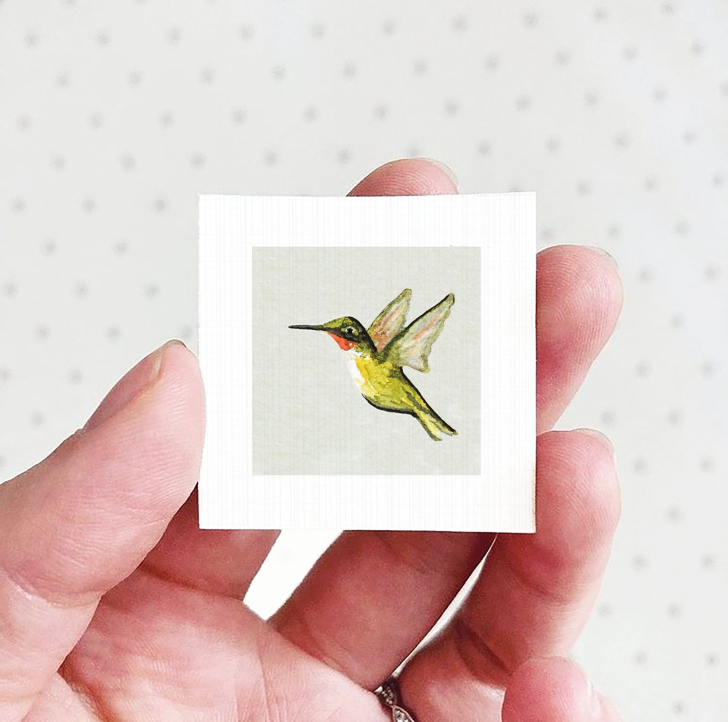 A hand holding a small square card with a painting of a hummingbird on it.