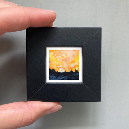 A hand is holding a small, square-shaped black frame containing a miniature painting of a sunset landscape.