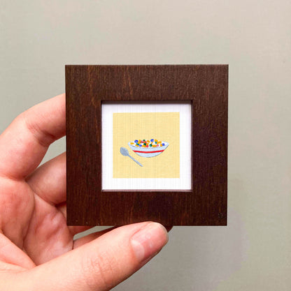A hand is holding a small square picture frame containing an illustration of a bowl of cereal.