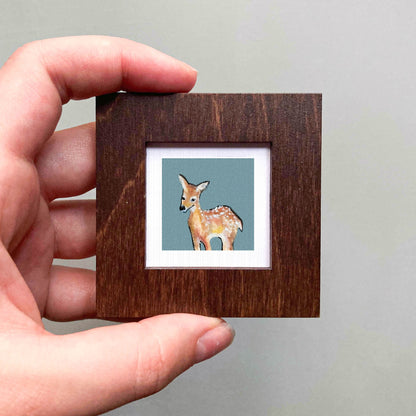 A hand holding a small wooden frame containing a painting of a deer.