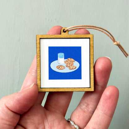 A person's hand is holding a small wooden frame containing an illustration of a plate with cookies and a glass of milk.