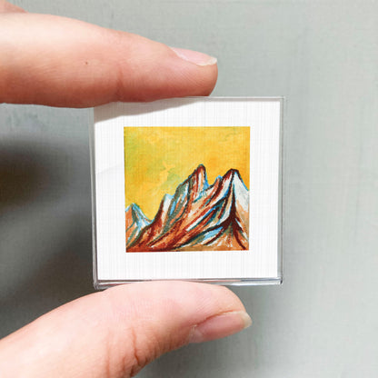 A person's hand is holding a small square picture of a mountain landscape with vibrant colors.
