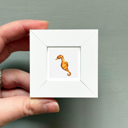 A hand is holding a small white square frame containing a miniature painting of a seahorse.