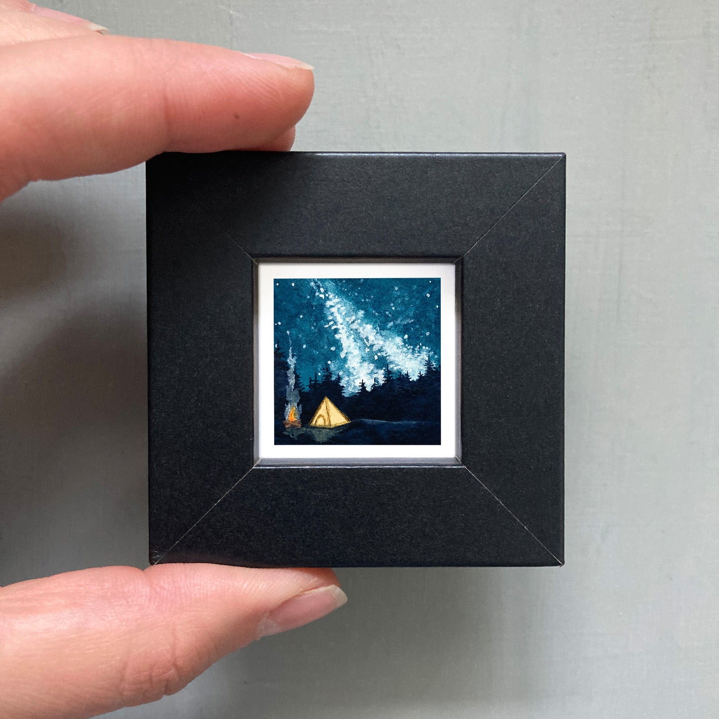 A person's hand is holding a small black frame containing a framed painting of a starry night sky with a tent in the foreground.