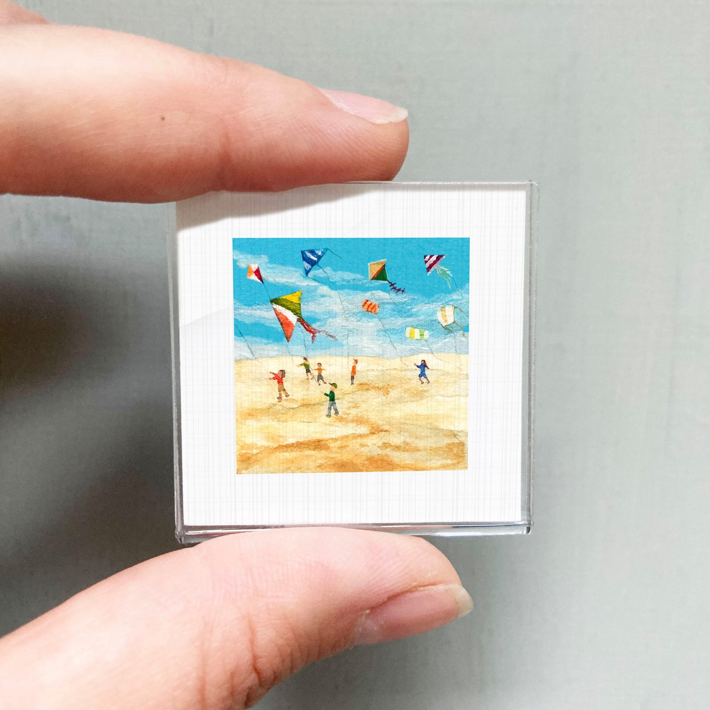 A person's hand is holding a small square picture depicting a beach scene with people flying colorful kites.