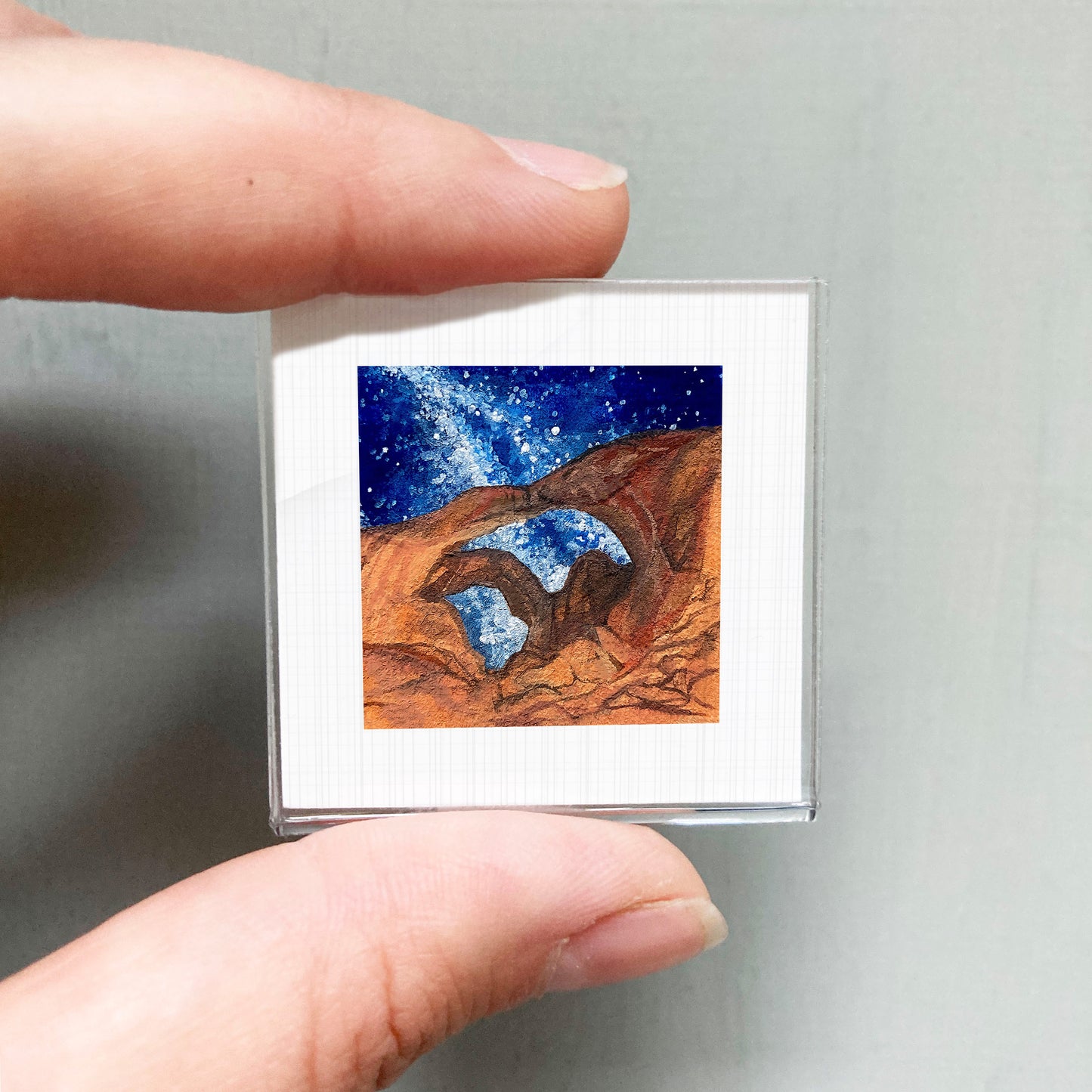A person's hand holding a small square picture of a desert landscape with a starry night sky.