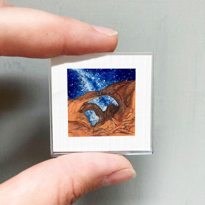 A person's hand holding a small square picture of a desert landscape with a starry night sky.