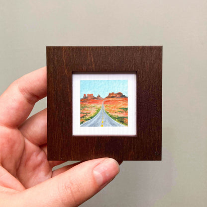 A hand is holding a small wooden frame containing a miniature painting of a desert landscape with a winding road leading to distant mountains.