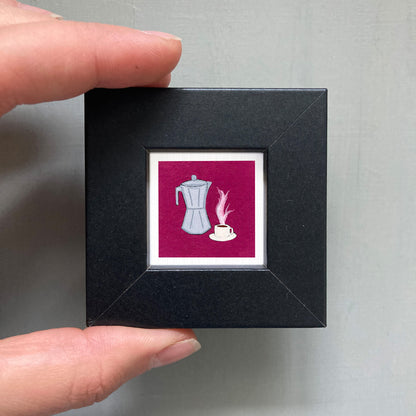 A hand is holding a small black square frame containing an illustration of a coffee maker and a cup of coffee.