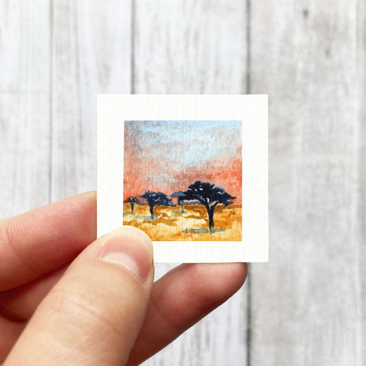A hand is holding a small square painting of a landscape with trees and a sky background.