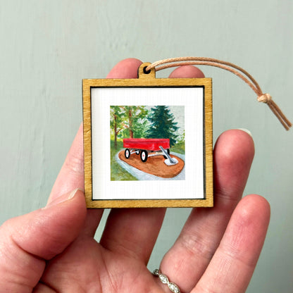 A person's hand holding a small wooden frame containing a painting of a red wagon on a dirt road surrounded by trees.