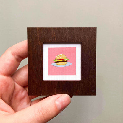 A hand is holding a small wooden frame containing a framed illustration of a hamburger.