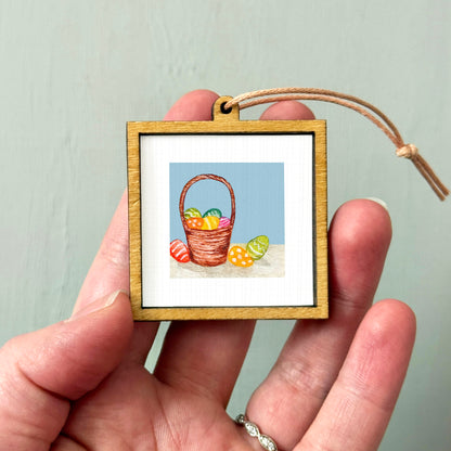 A person's hand holding a small wooden frame containing a painting of a basket filled with colorful Easter eggs.