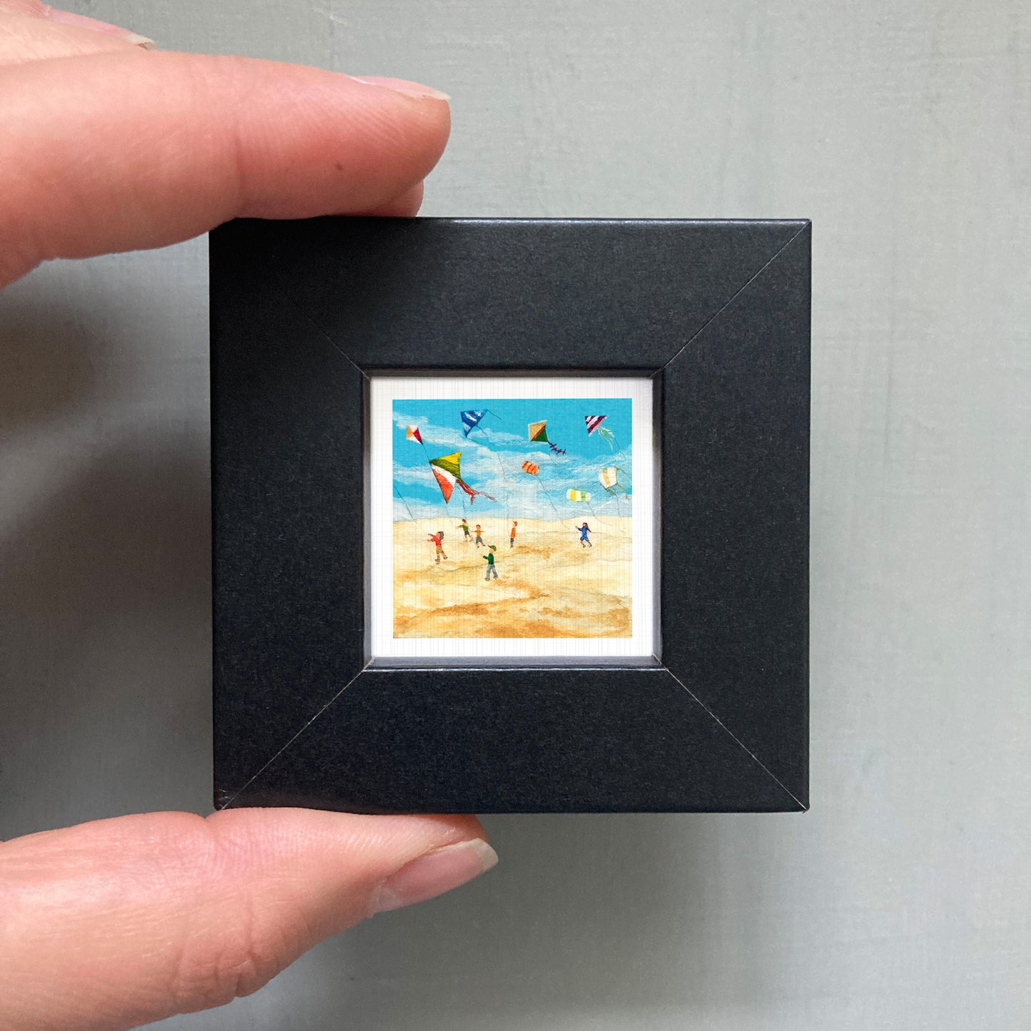 A person's hand is holding a small black frame containing a framed painting of people flying kites on a beach.
