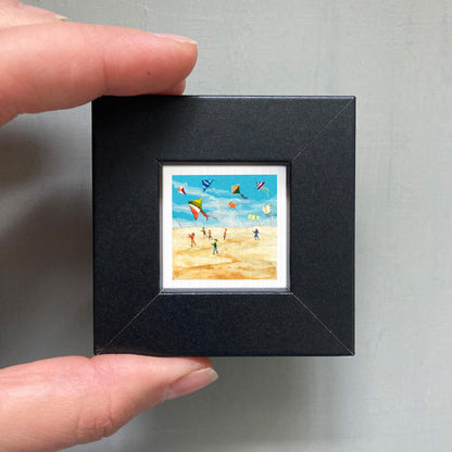 A person's hand is holding a small black frame containing a framed painting of people flying kites on a beach.