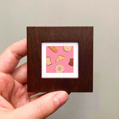 A hand is holding a small square frame containing an illustration of various pastries, including a croissant, a donut, and a muffin.