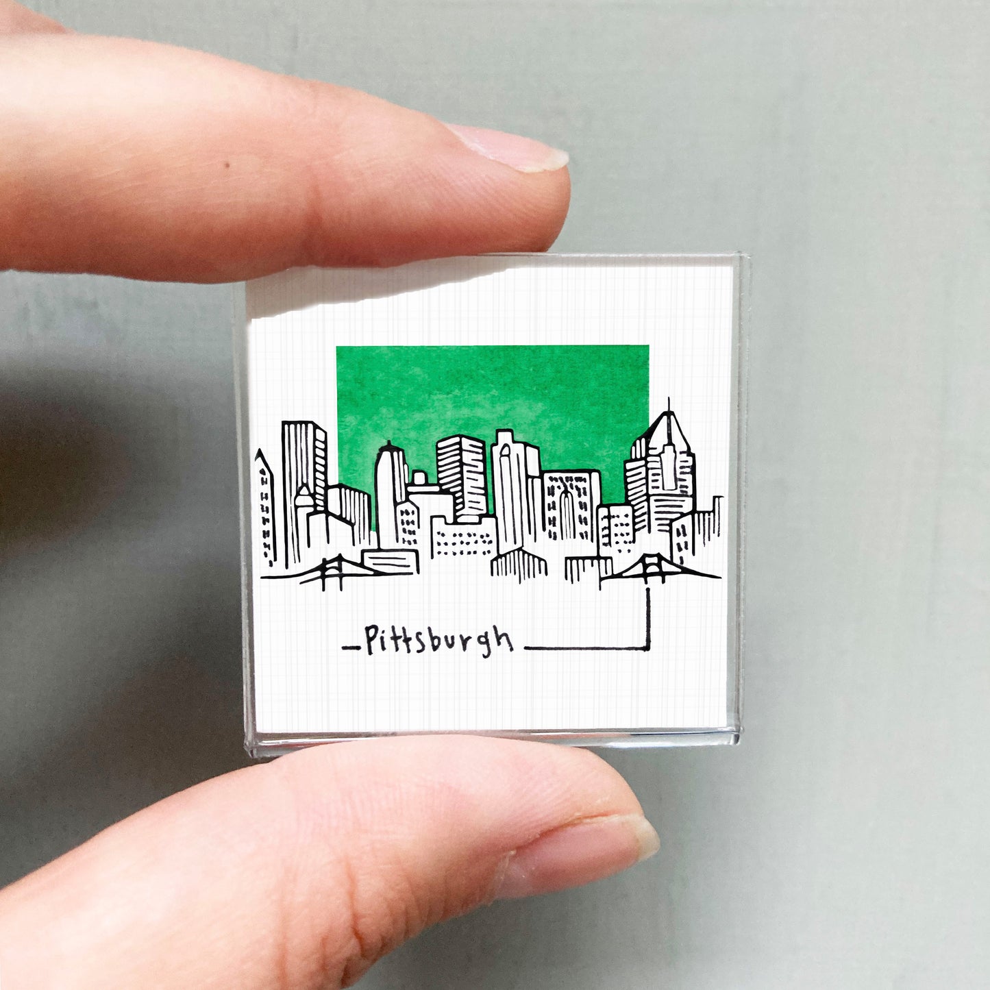 A hand holding a small square magnet with a drawing of a city skyline and the word "Pittsburgh" written below it.