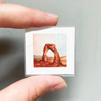 A person's hand holding a small square picture of the Delicate Arch, a natural geological formation in the Arches National Park in Utah.