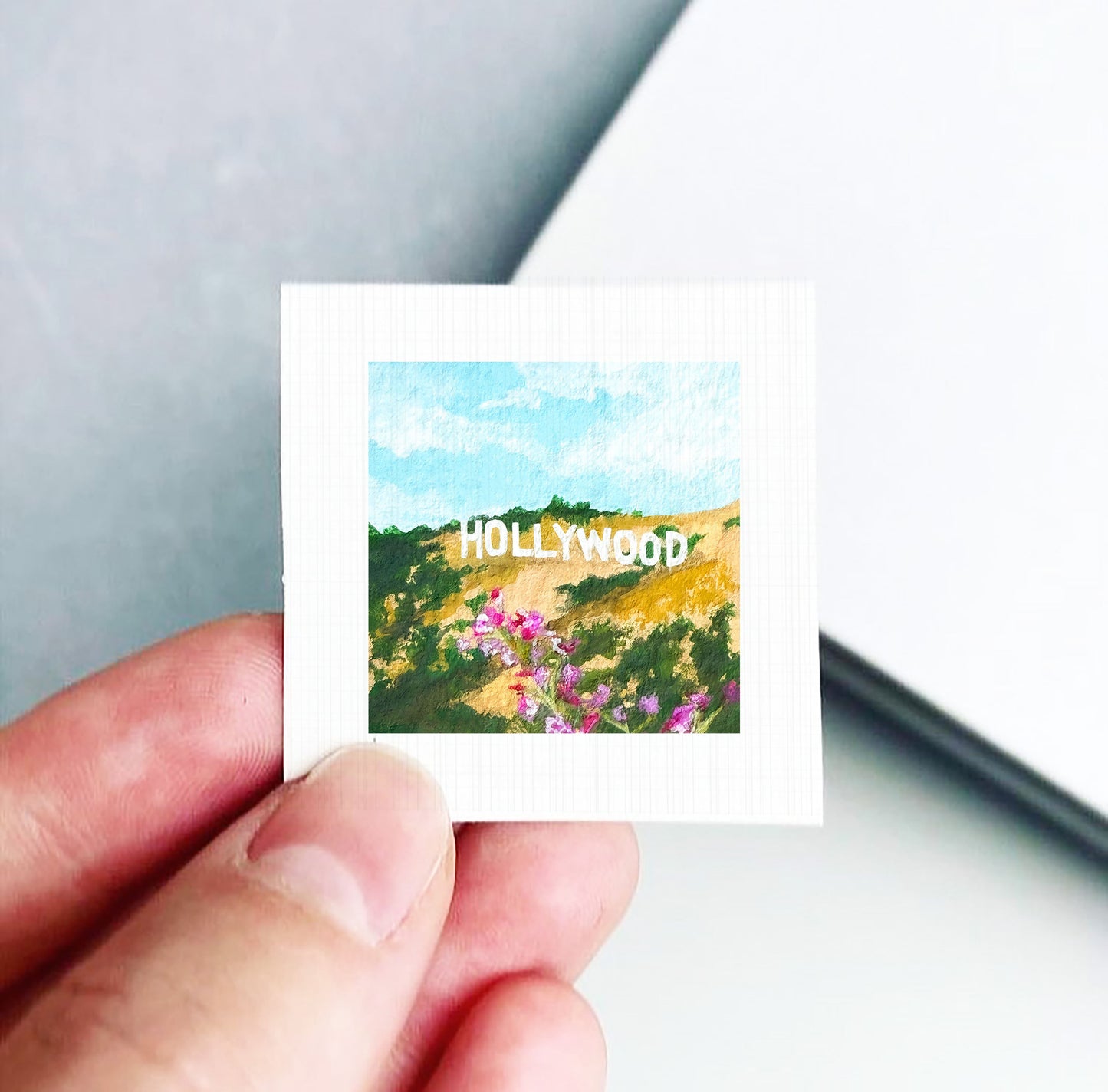 A hand holding a small square-shaped piece of paper with a painting of a scenic landscape featuring the Hollywood sign in the background.
