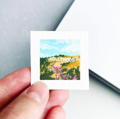 A hand holding a small square-shaped piece of paper with a painting of a scenic landscape featuring the Hollywood sign in the background.