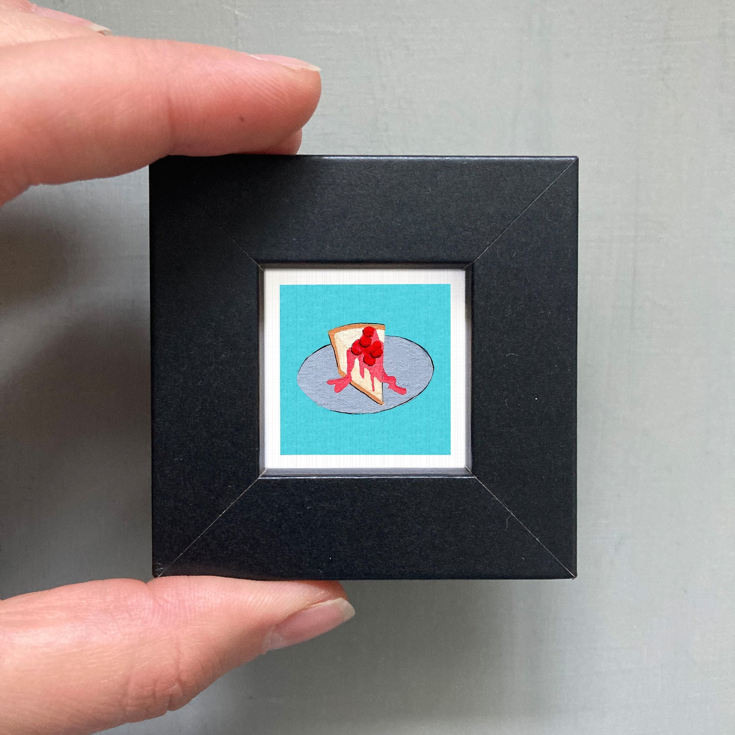 A hand is holding a small black frame containing a framed image of a slice of cake with red frosting.