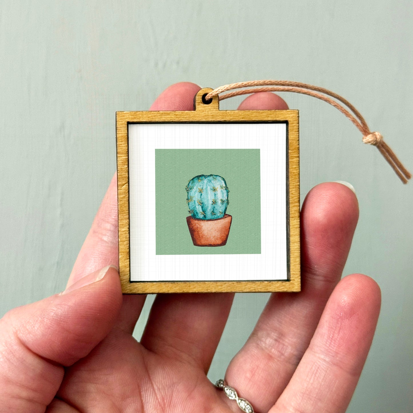 A hand holding a small wooden frame containing a framed illustration of a cactus plant.